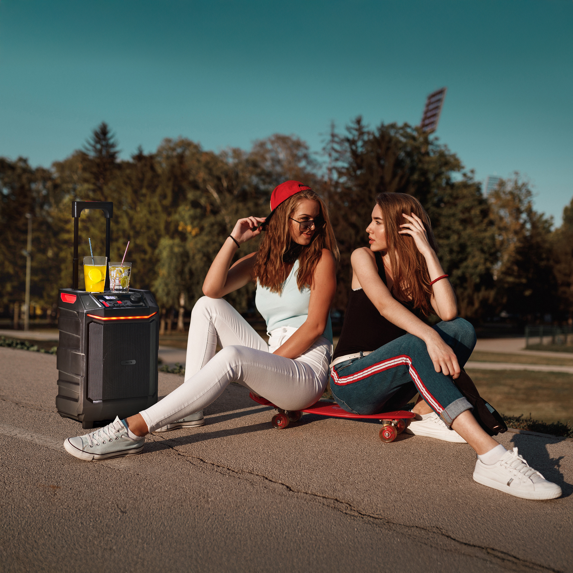 Dos mujeres sentadas en monopatín. Altavoz y bebidas cerca. Árboles y cielo azul al fondo.