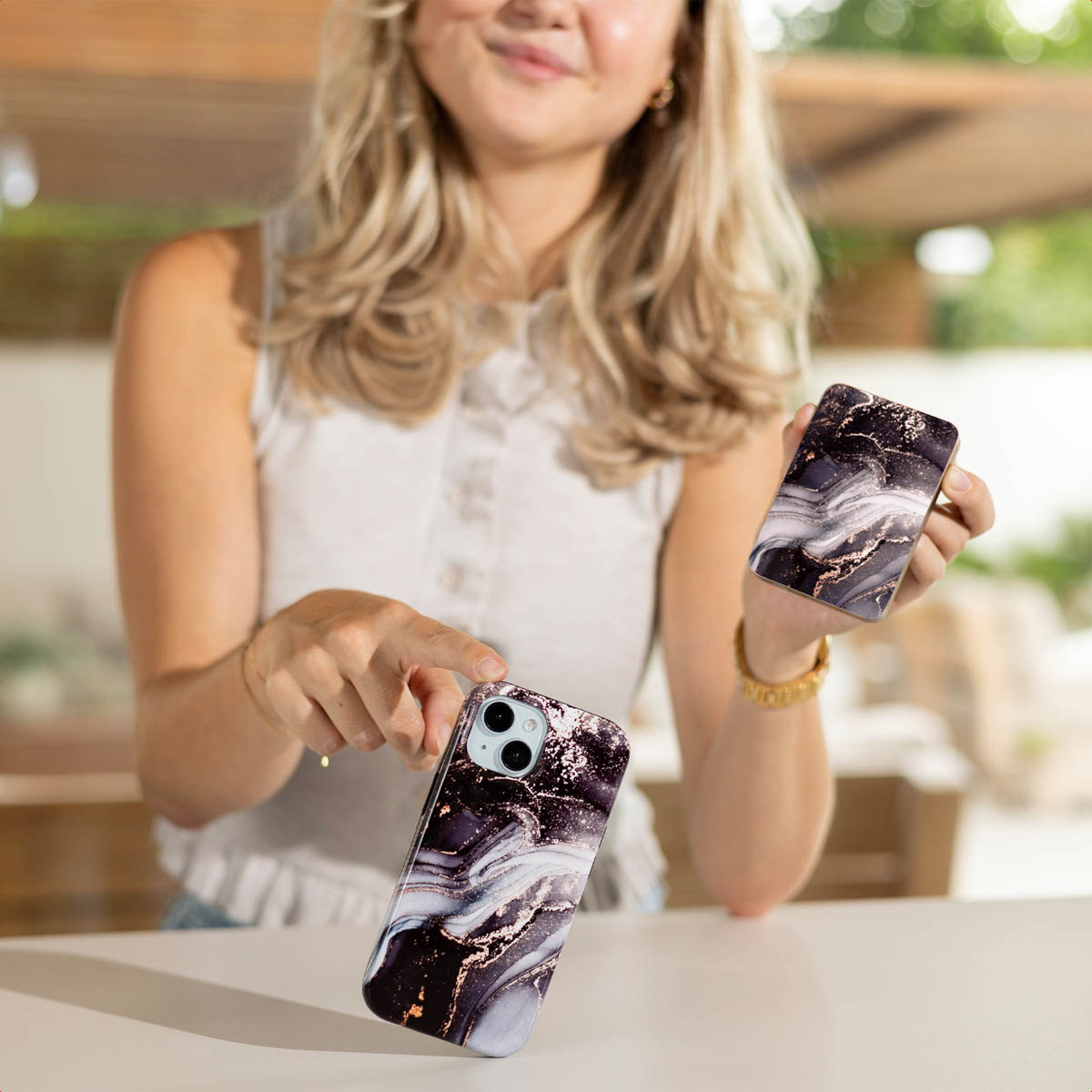 Een vrouw houdt twee telefoonhoesjes vast met een marmerpatroon. Eén valt. Ze glimlacht en draagt een witte top en gouden sieraden.