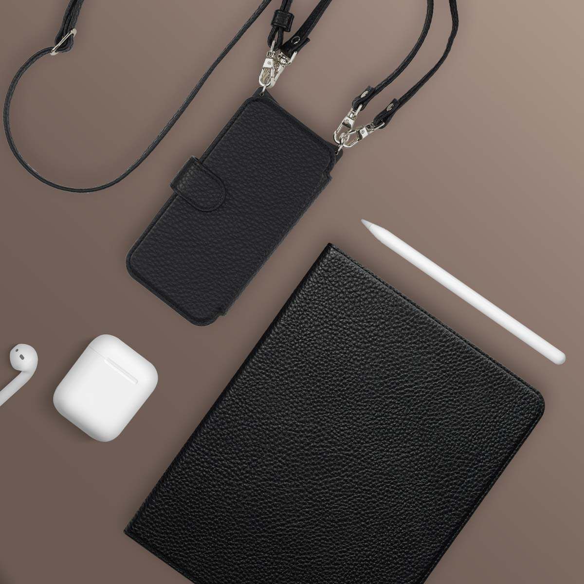 Zwarte telefoonhoes met riem, een notitieboek, stylus en AirPods op een bruin oppervlak. Items zijn van boven naar beneden gerangschikt.