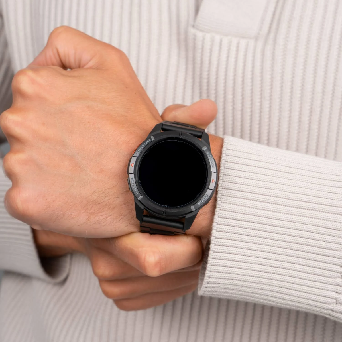 Osoba nosząca czarny smartwatch z pustym ekranem. Zegarek jest na jej lewym nadgarstku, z jasnym swetrem w tle.