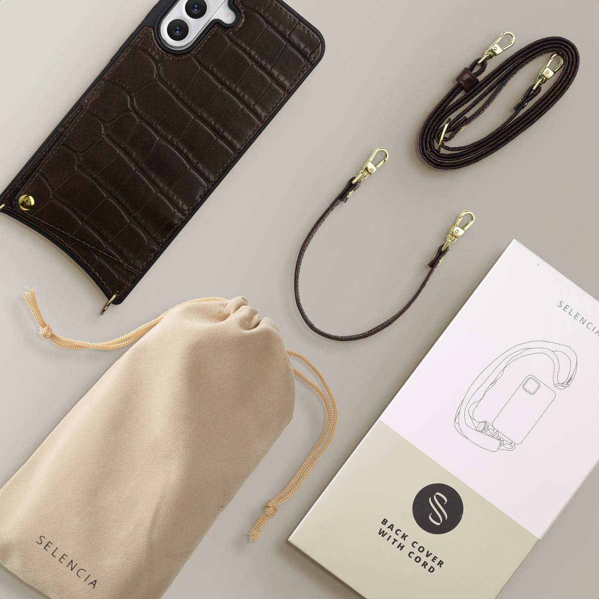 Una custodia per telefono, un cinturino, una custodia e un imballaggio sono disposti su una superficie grigio chiaro. La custodia del telefono è marrone con un motivo a coccodrillo.