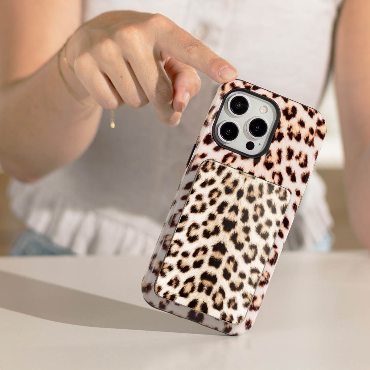 Een hand raakt een telefoonhoesje met luipaardprint en ingebouwde grip aan. De telefoon ligt op een wit oppervlak.