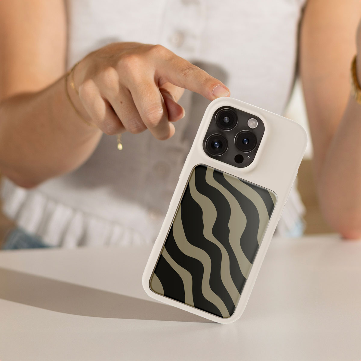 Een hand houdt een witte telefoonhoes met een zebrapatroon vast. Een persoon draagt een wit overhemd en gouden armbanden. De telefoon ligt op een witte tafel.