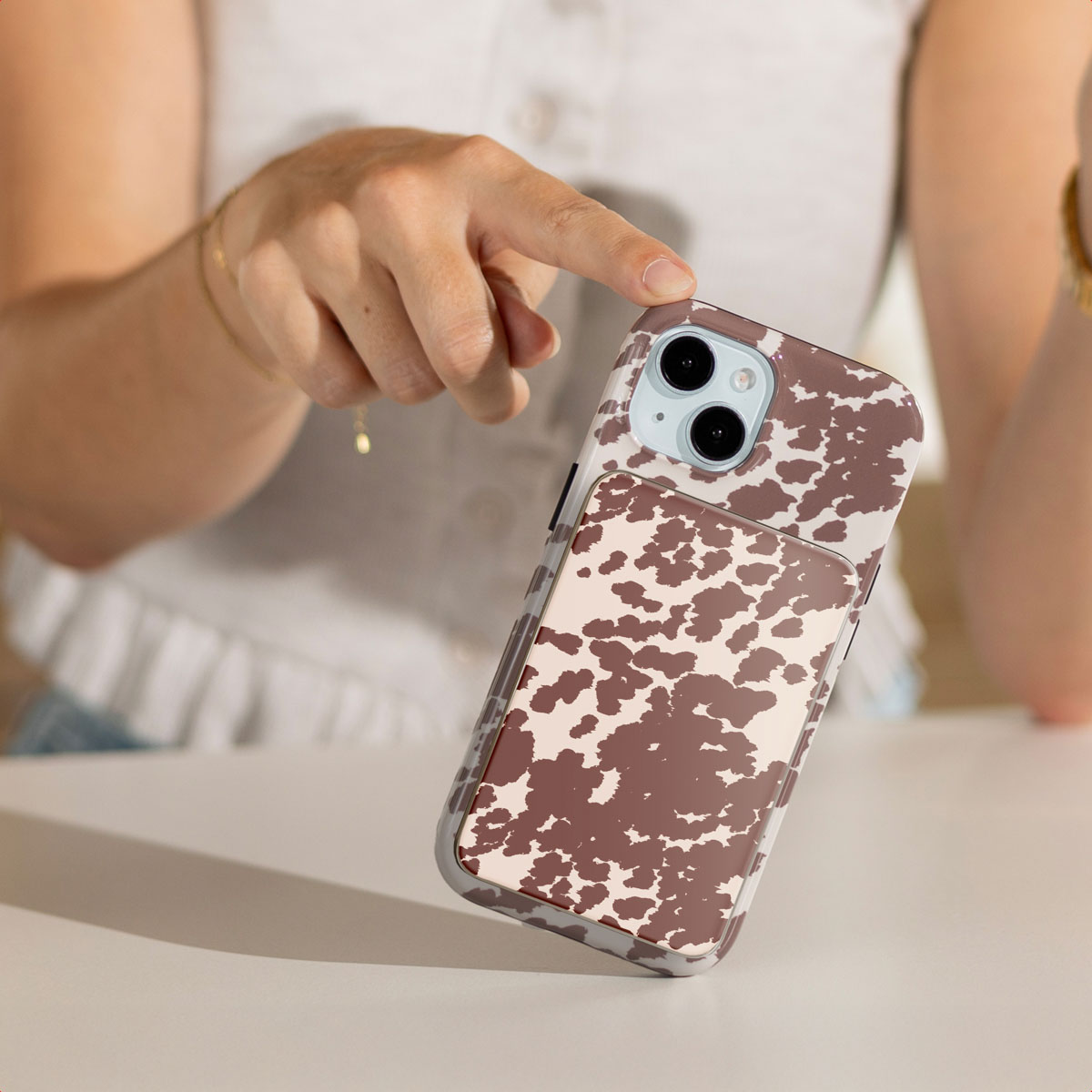 Een hand die een telefoon vasthoudt. De telefoon heeft een bruin-witte koeienprint hoes. De achtergrond is een witte tafel.