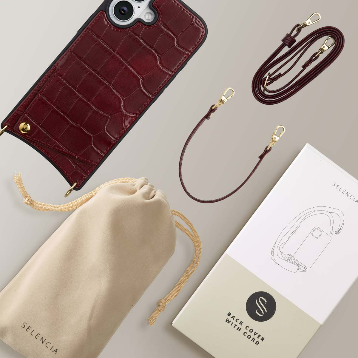 Een bordeauxrode telefoonhoes, riem, etui en verpakking worden getoond op een lichtgrijze ondergrond. Accessoires met gouden hardware.