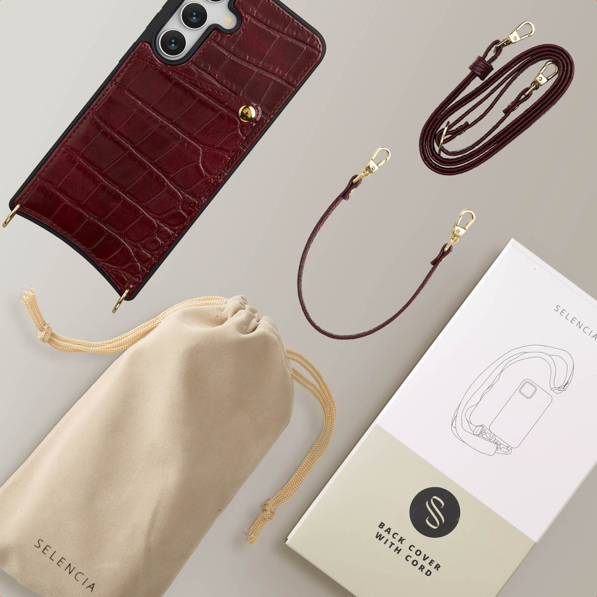 Een telefoonhoesje en accessoires, waaronder een band en een etui. De hoes is bordeauxrood met gouden details. Verpakking op de achtergrond.