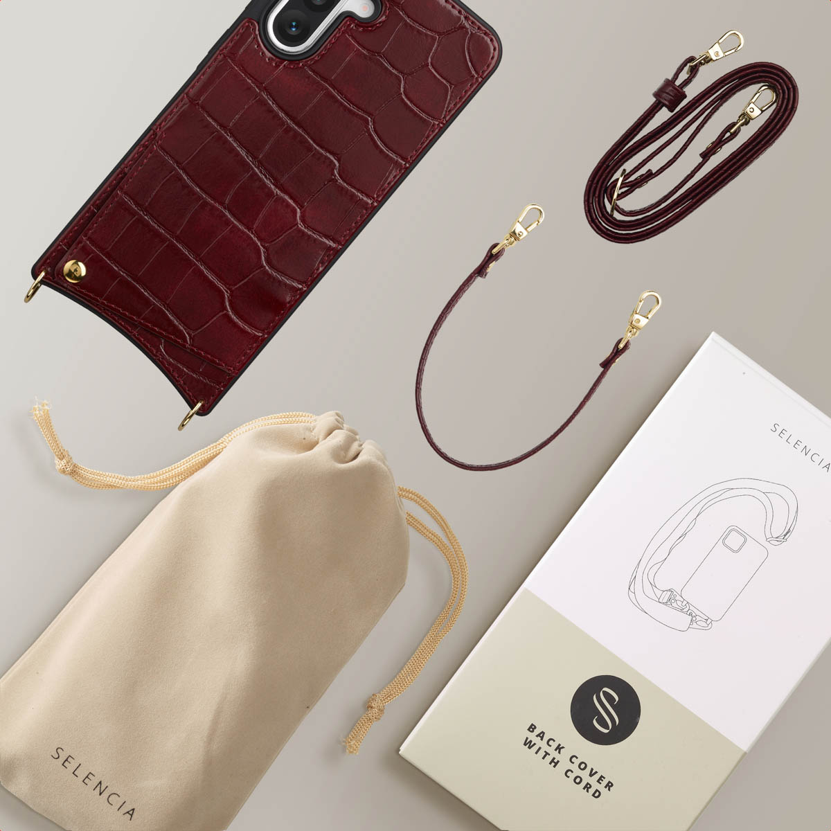 Een telefoonhoesje en accessoires, waaronder een riem en een tas, liggen op een licht oppervlak. De hoes is bordeauxrood, met een textuur.