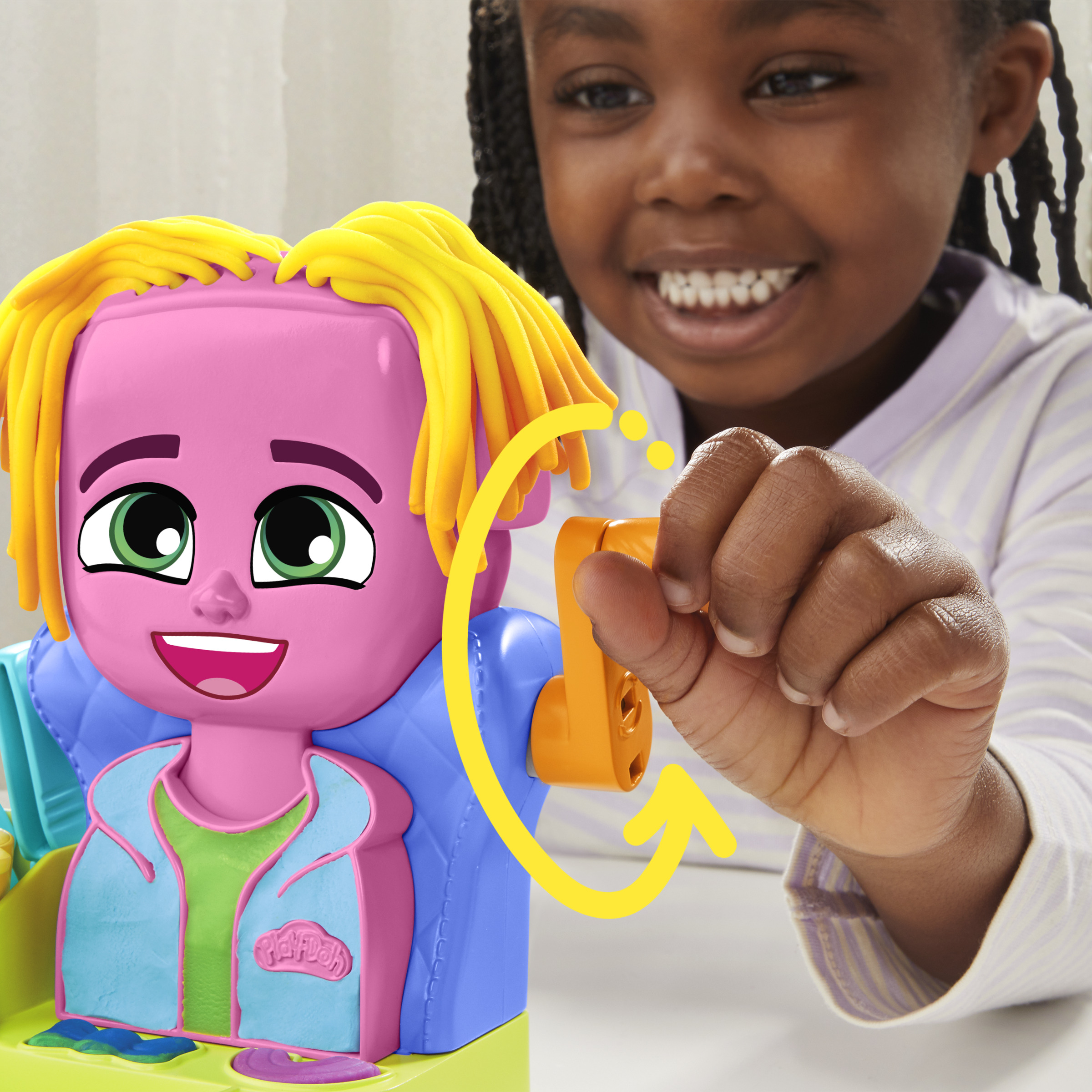 Una niña sonríe a un juguete. El juguete es rosa con pelo amarillo, y la niña sostiene una palanca del juguete.