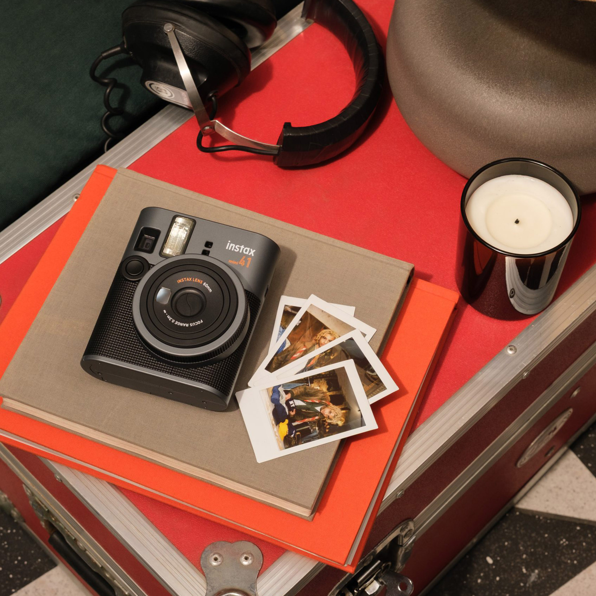 Gri bir instax kamera, fotoğraflar, kulaklıklar ve bir mum kırmızı bir bavulun üzerinde duruyor. Bir kitap ve turuncu kağıt da var.