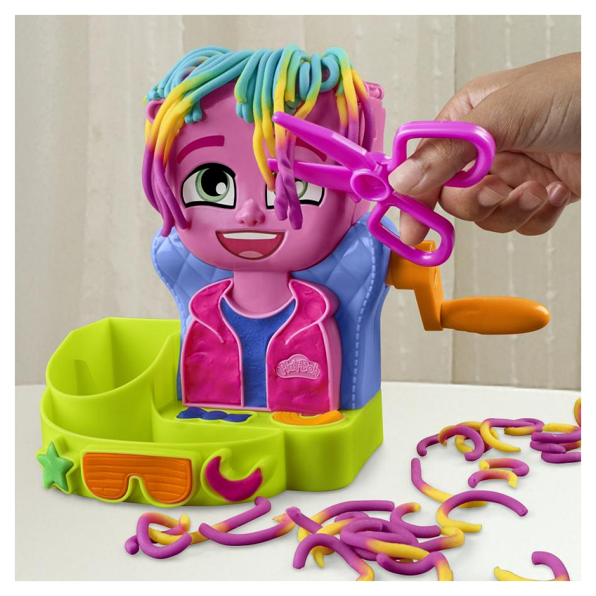 Un juguete Play-Doh rosa con pelo y tijeras. Una mano sostiene las tijeras cerca del pelo. Cadenas coloridas de Play-Doh están sobre la superficie.