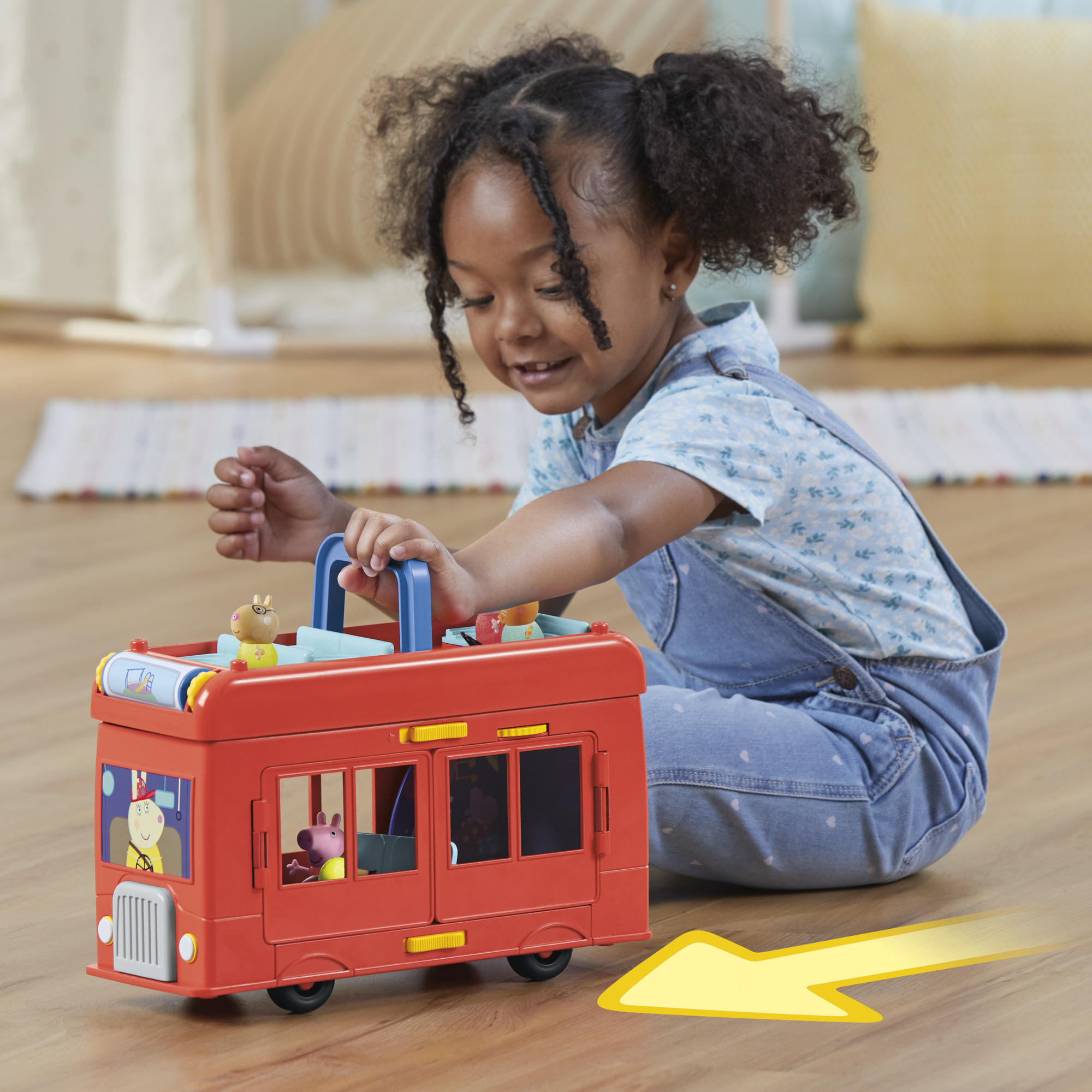 Una niña juega con un autobús de juguete rojo. Sonríe, sujetando el asa. El autobús tiene figuras de animales dentro.