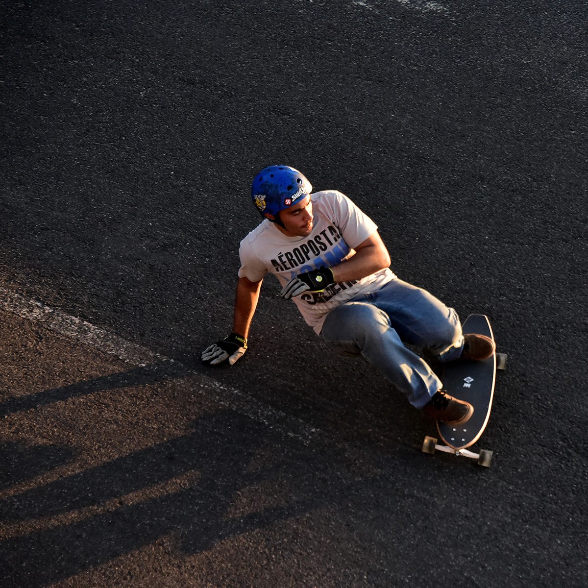 Ein Mann mit blauem Helm und Handschuhen fährt auf Asphalt Skateboard. Er beugt sich nach unten. Das Board ist schwarz.