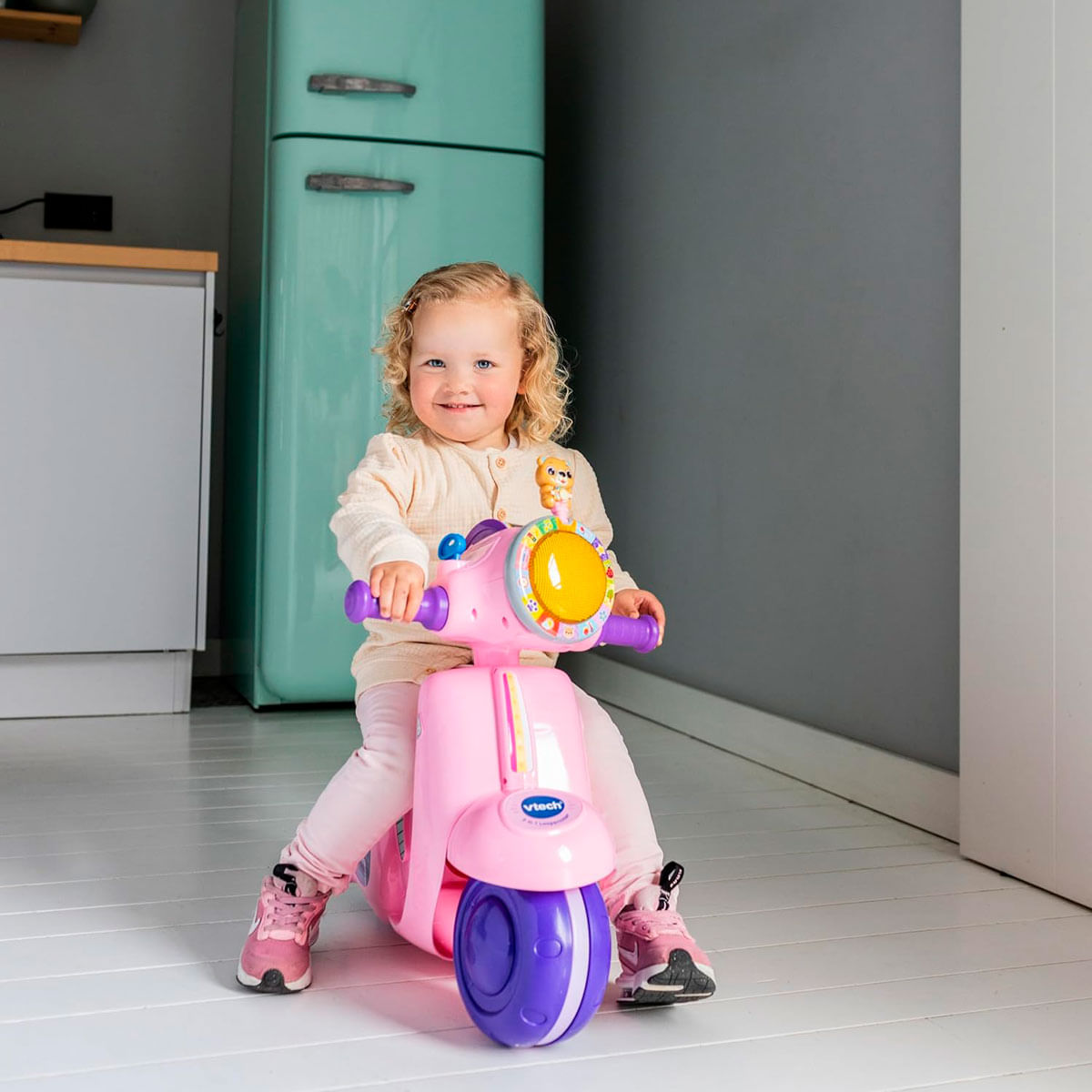 Una niña sonriente está sentada en un scooter de juguete rosa. Detrás de ella hay un refrigerador verde claro. El scooter tiene una luz amarilla y ruedas moradas.