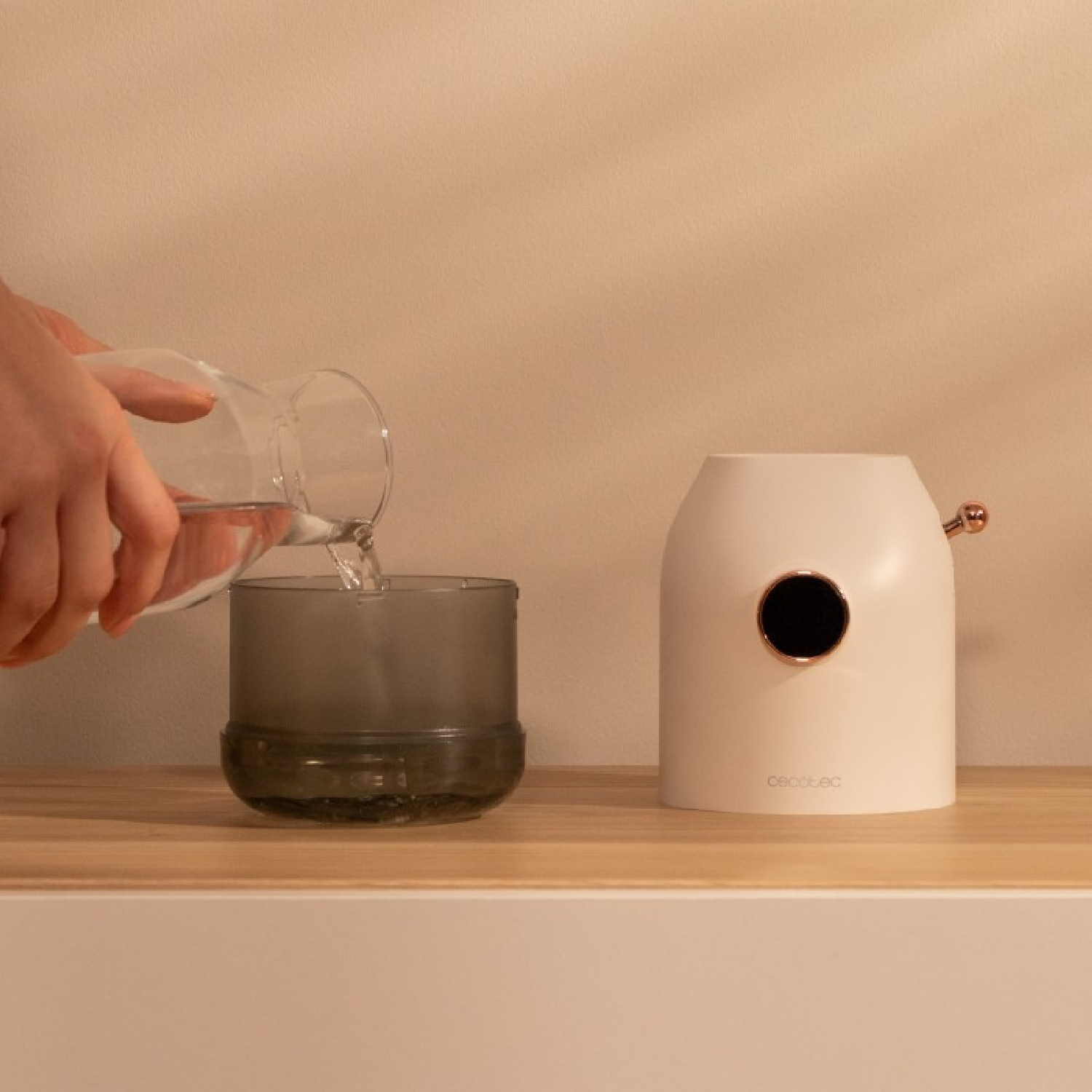 Eine Hand gießt Wasser aus einem klaren Krug in ein graues Glas. Rechts steht ein weißer Diffusor mit goldenem Akzent.