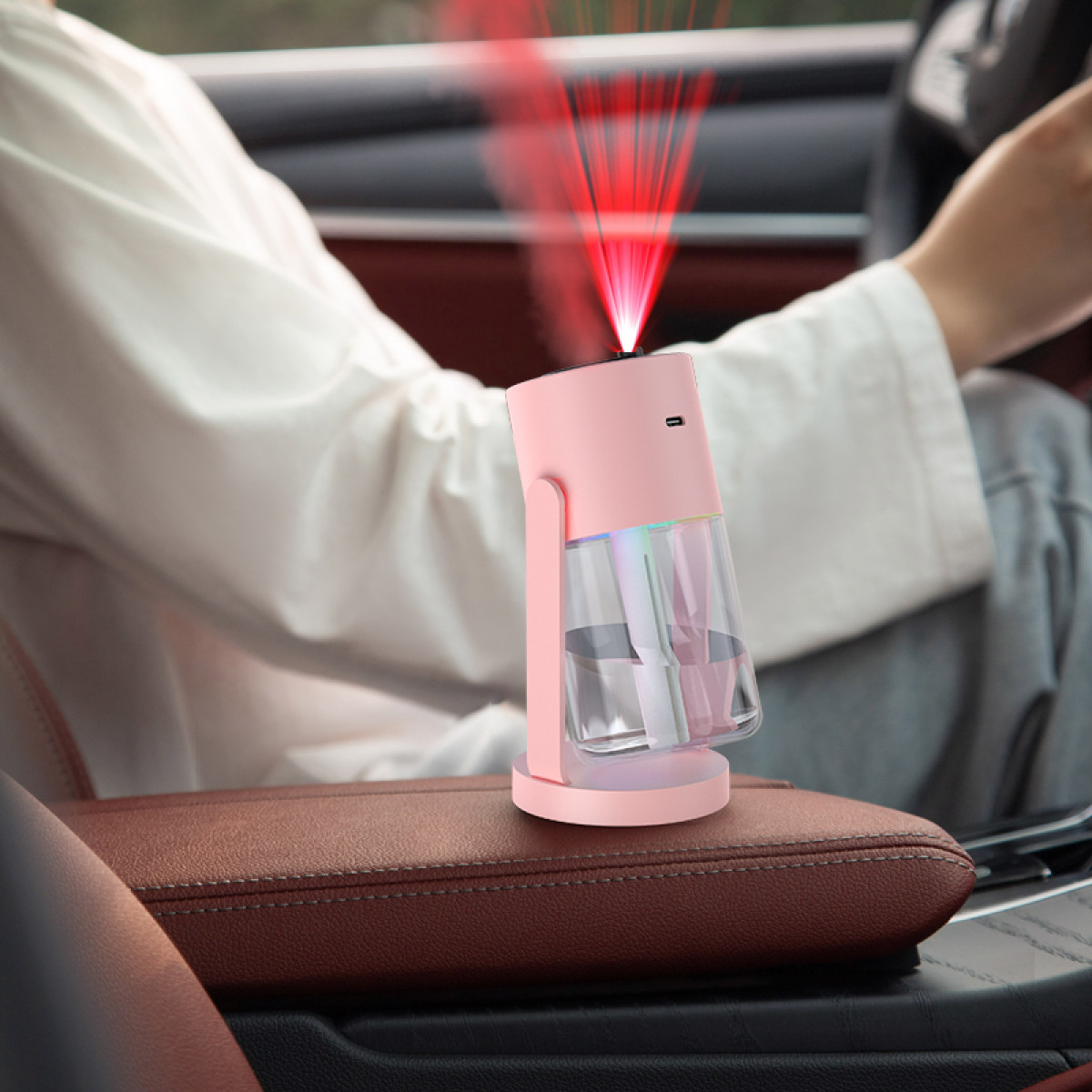 Ein rosa Auto-Lufterfrischer mit rotem Licht, in einem Auto. Das Gerät steht auf der braunen Armlehne. Eine Person fährt.