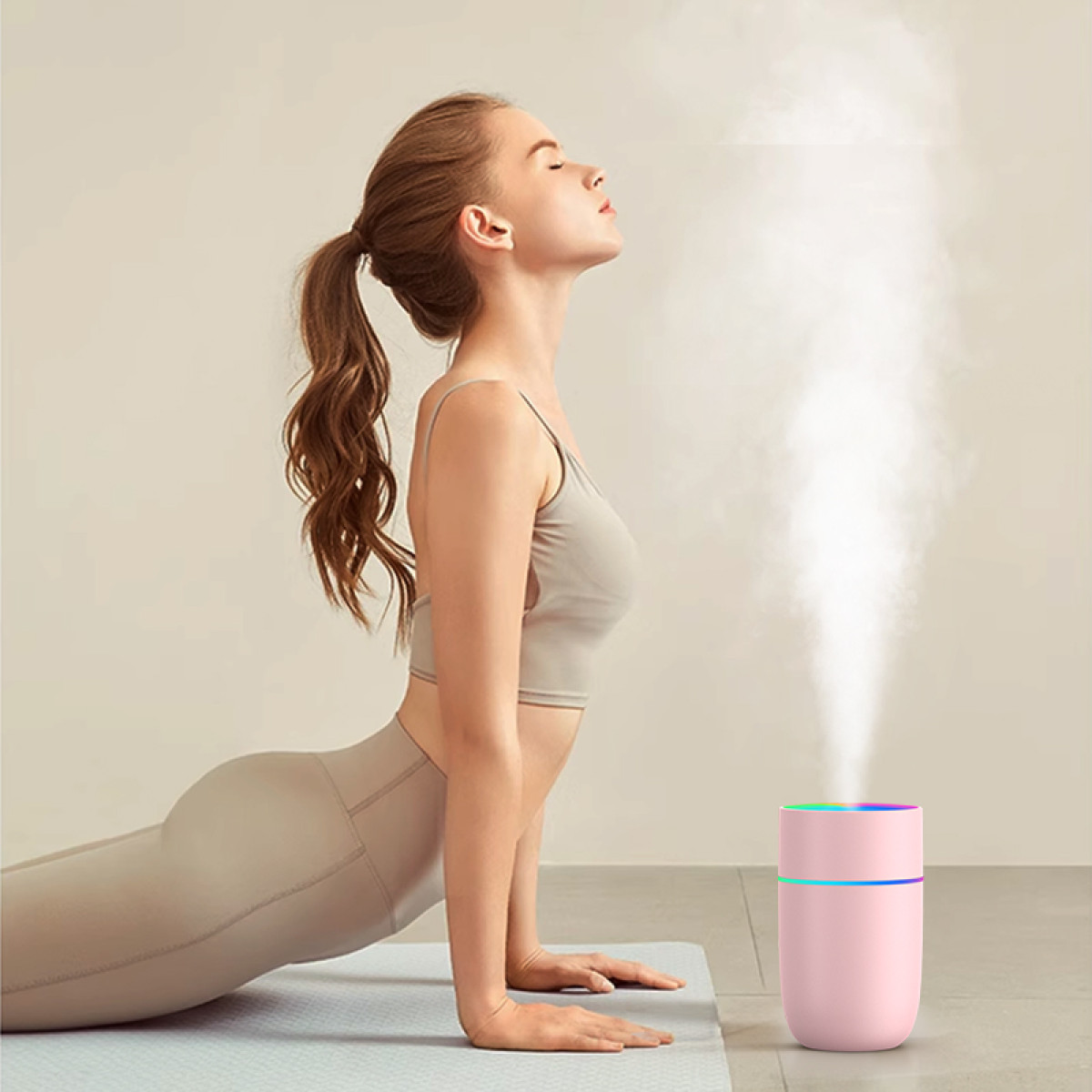 Eine Frau in Yoga-Pose, Augen geschlossen, neben einem rosa Luftbefeuchter, der Dampf abgibt. Neutraler Hintergrund. Sie trägt Sportkleidung.