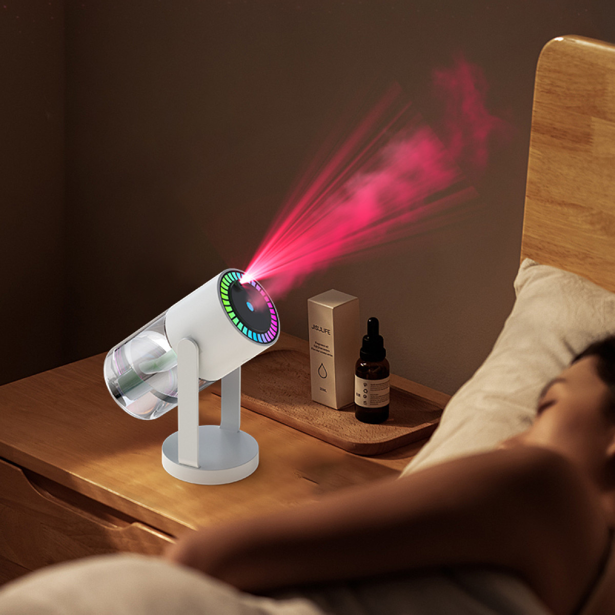 Ein weißer Projektor strahlt rotes Licht aus. Eine Person schläft im Bett. Eine Flasche und eine Schachtel stehen auf dem Nachttisch.