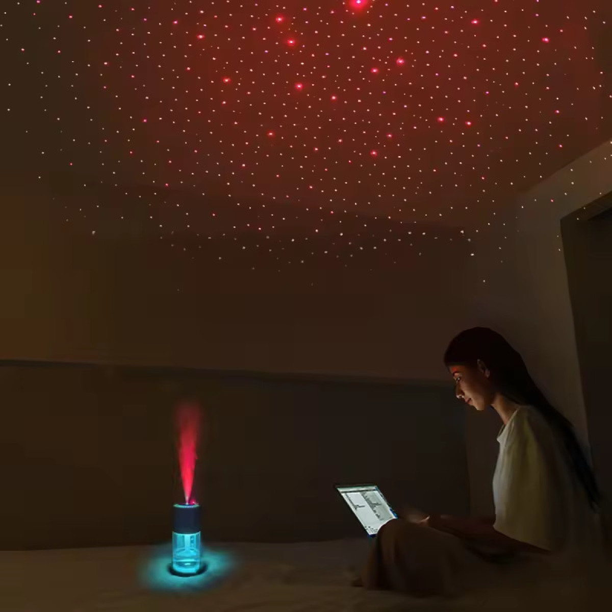 Een vrouw zit op een bed met een tablet, onder een plafond verlicht met rode sterren. Een apparaat zendt een rode straal uit.