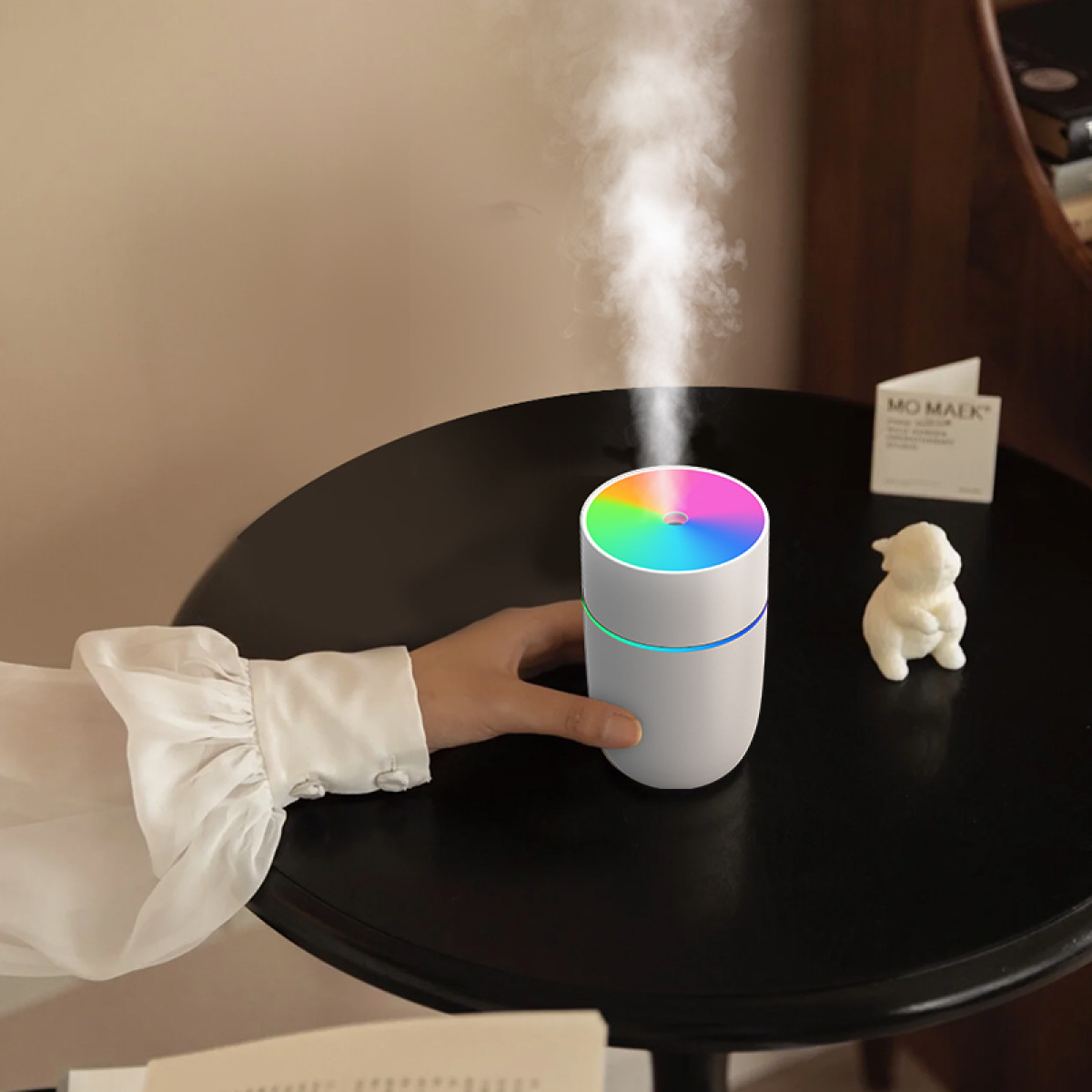 Ein weißer Luftbefeuchter mit Regenbogenlicht steht auf einem schwarzen Tisch. Eine Hand berührt ihn. Eine kleine Figur und eine Karte sind in der Nähe.