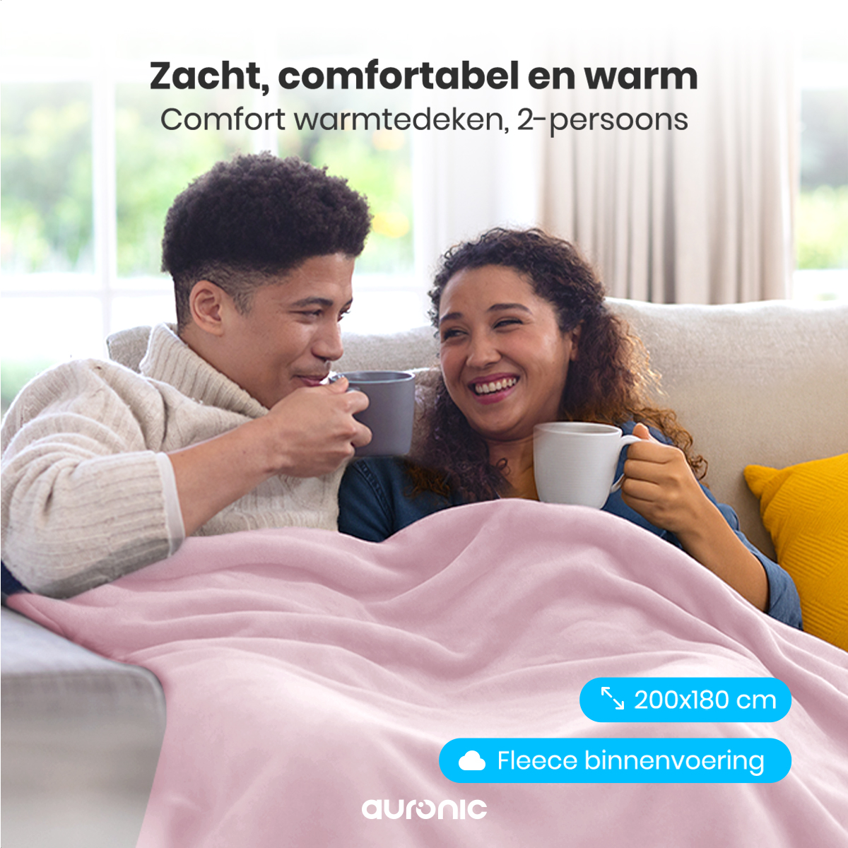 A couple on a sofa, covered by a pink blanket. They are smiling and holding mugs. Text says 'Comfort warmtedeken, 2-persoons'.