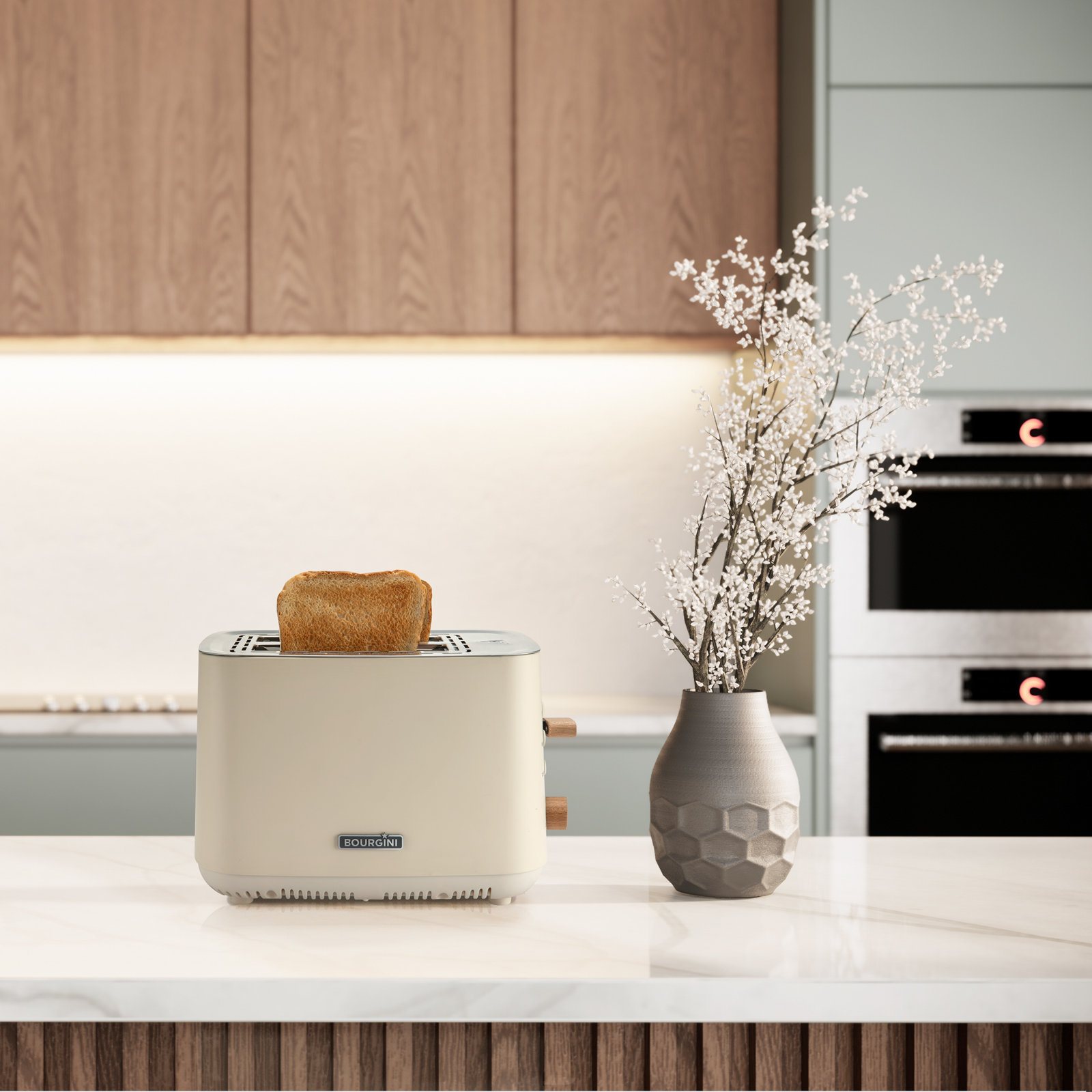 Een beige broodrooster met toast staat op een wit aanrecht. Een vaas met witte bloemen rechts. Keuken op de achtergrond.