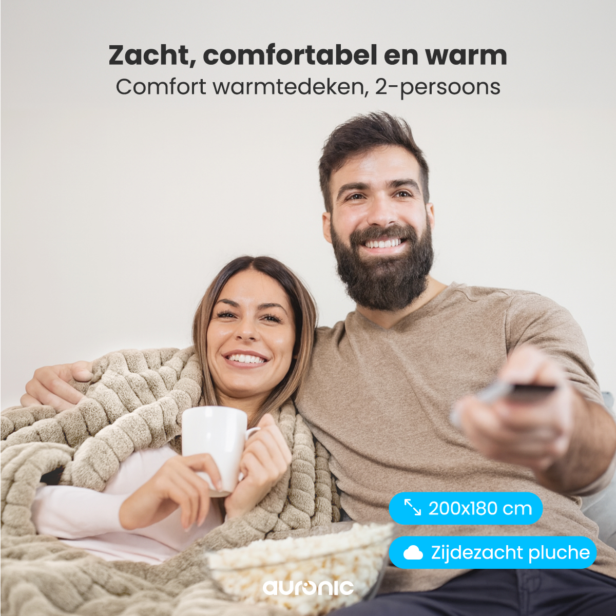 A couple smiles, wrapped in a blanket, holding a mug and remote. There is popcorn and text that says 'Zacht, comfortabel en warm.'