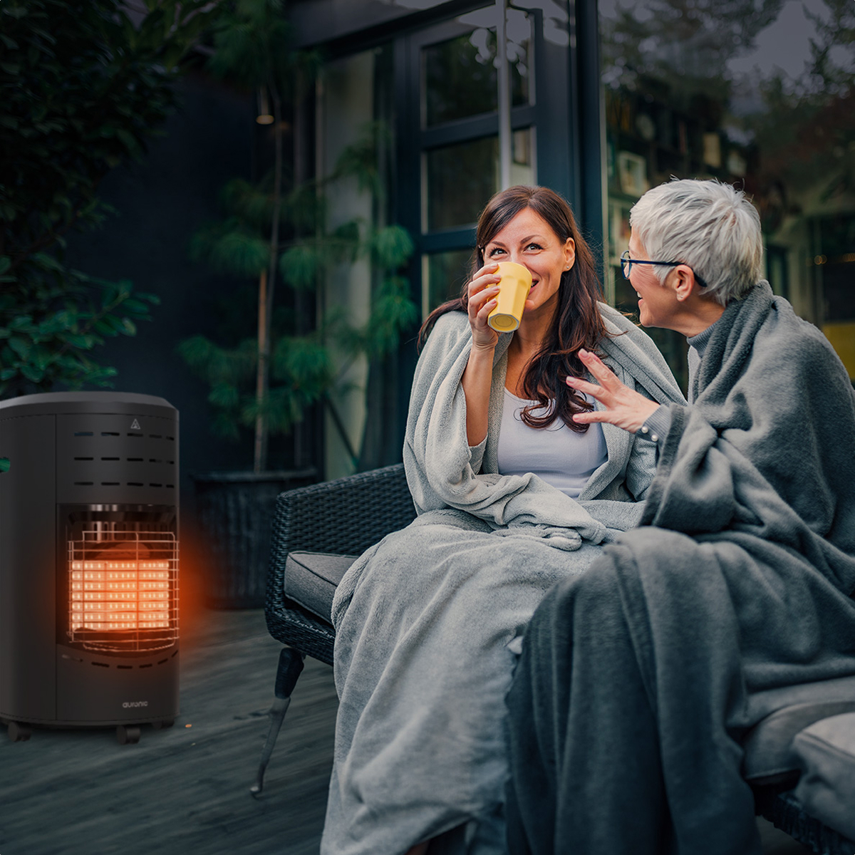 Twee vrouwen op een terras, gehuld in dekens. Eén drinkt uit een gele mok. Een zwarte kachel gloeit in de buurt.