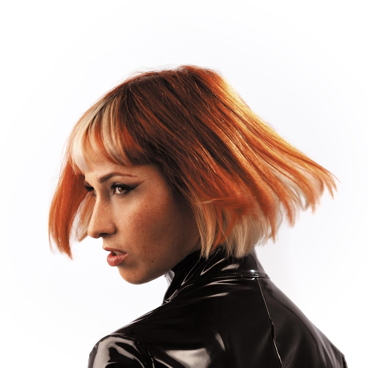 Una donna con capelli rossi e biondi guarda a sinistra. Indossa un top nero lucido su sfondo bianco.