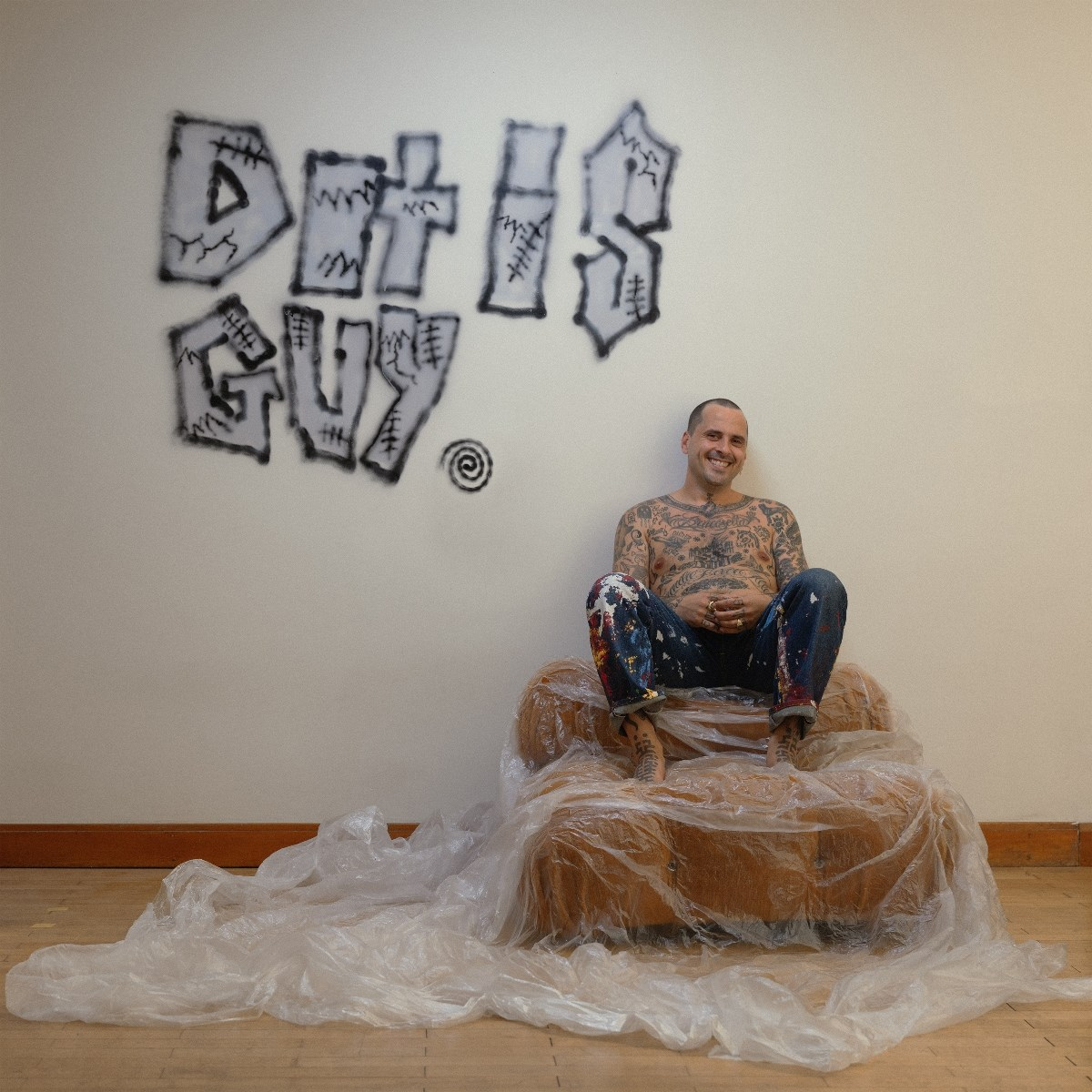 Een getatoeëerde man zit op een ingepakte stoel. Graffiti op de muur: 'Death is Guy'. Hij lacht naar de camera.