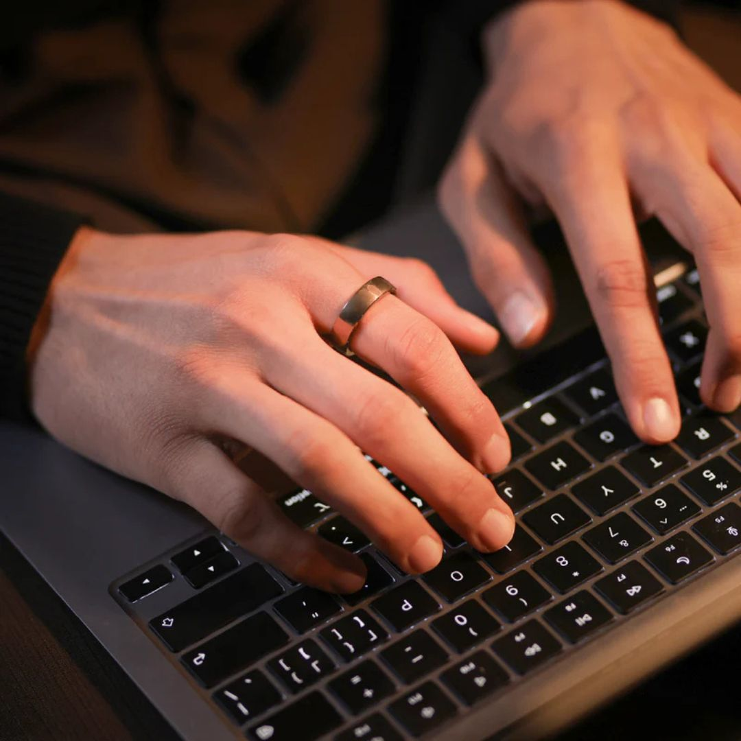 Hände tippen auf einer Laptop-Tastatur. Ein goldener Ring ist am Finger. Der Hintergrund ist verschwommen, dunkler Pullover.
