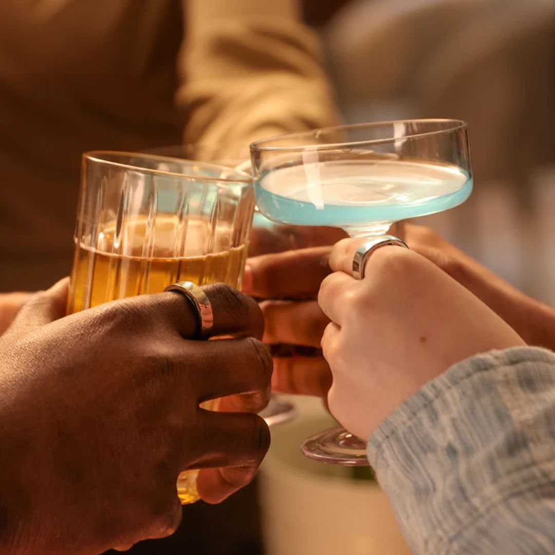 Hände stoßen mit Getränken an. Ein Glas hat bernsteinfarbene Flüssigkeit, das andere blau. Ringe sind an Fingern.