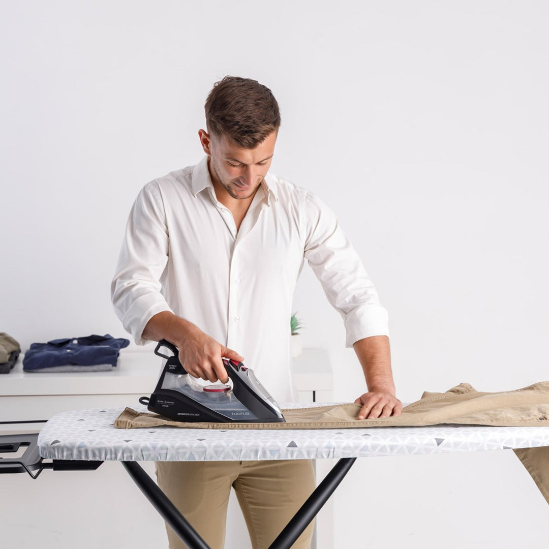 Mężczyzna w białej koszuli prasuje spodnie na desce do prasowania. Inne ubrania leżą na stole. Białe tło.