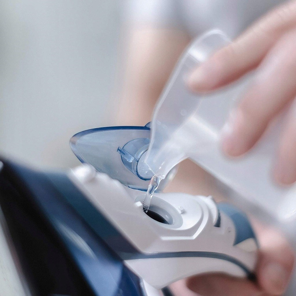 Osoba wlewa wodę z plastikowego pojemnika do żelazka. Żelazko jest biało-niebieskie. Tło jest rozmyte.