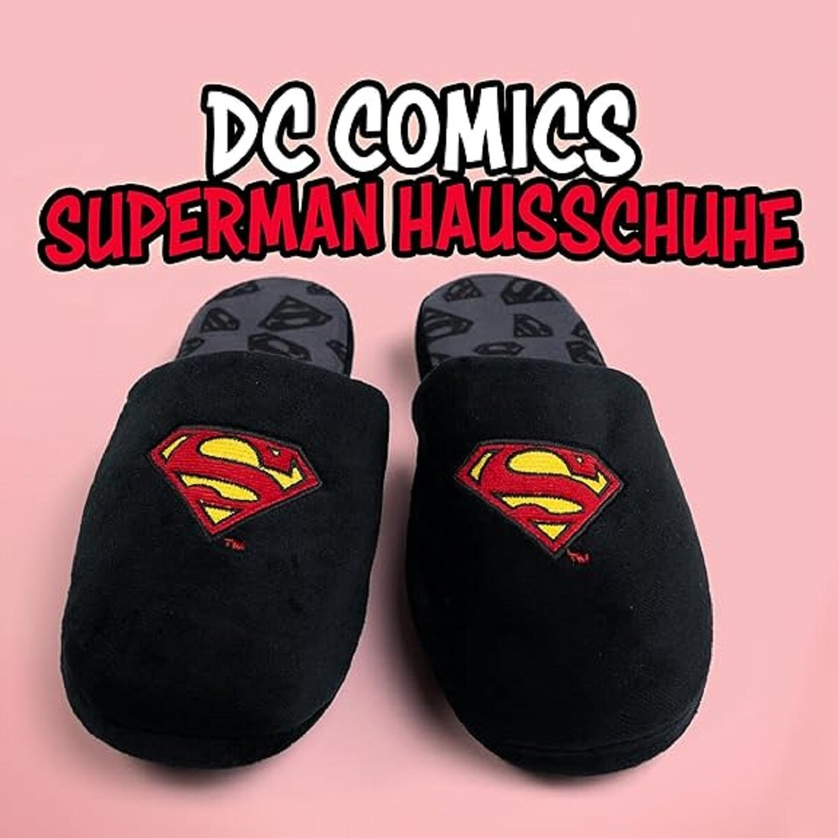 Paar schwarze Superman-Hausschuhe mit dem Superman-Logo auf der Vorderseite, vor rosa Hintergrund.