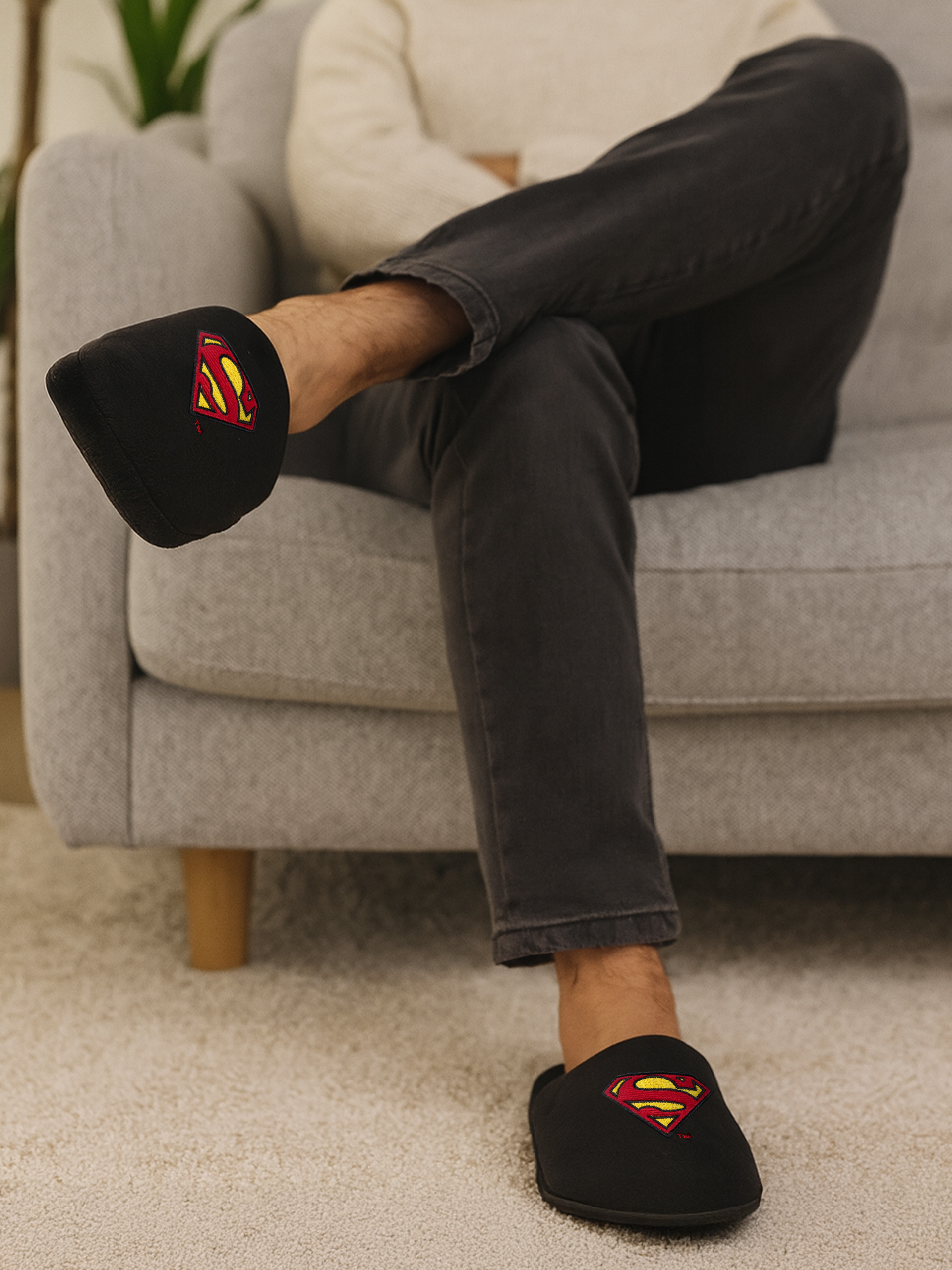 Person trägt schwarze Superman-Hausschuhe und sitzt auf einem grauen Sofa. Die Hausschuhe haben das Superman-Logo.