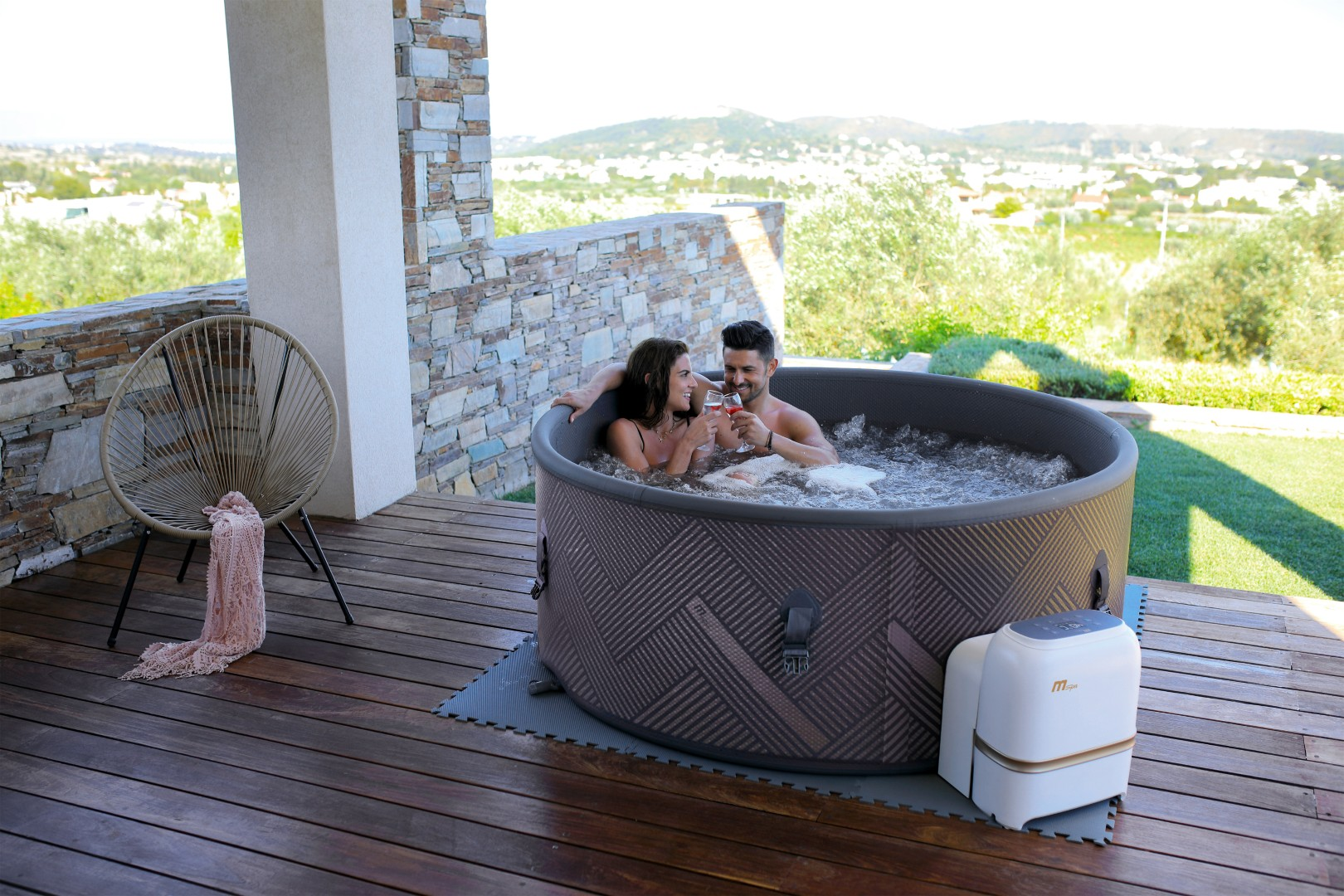 Ein Paar stößt in einem Whirlpool mit Wein an. Ein Stuhl und eine Decke stehen in der Nähe. Im Hintergrund ist eine Landschaft zu sehen.
