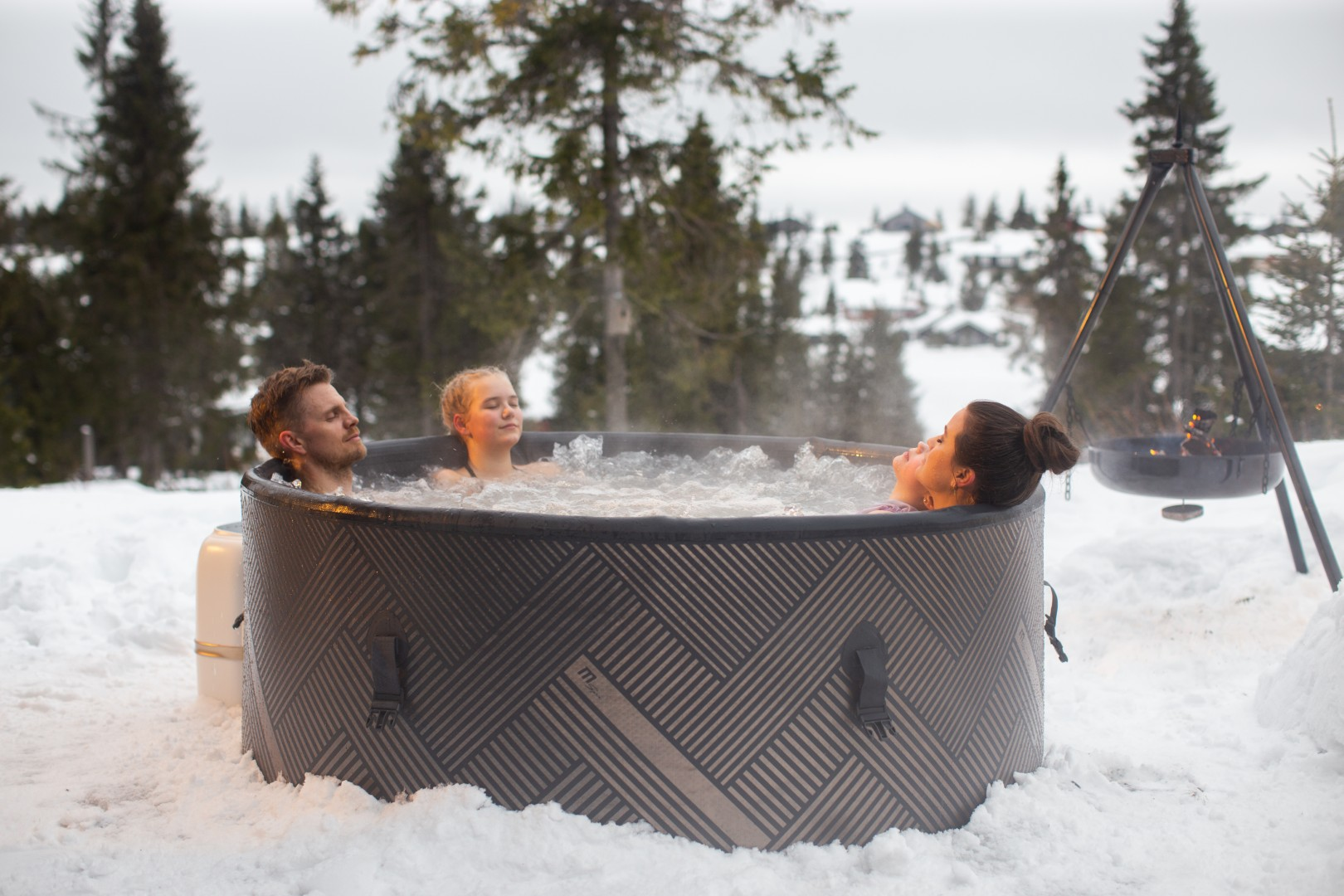 Drei Personen entspannen sich in einem Whirlpool im Freien, umgeben von Schnee. Bäume und eine Feuerstelle sind im Hintergrund.
