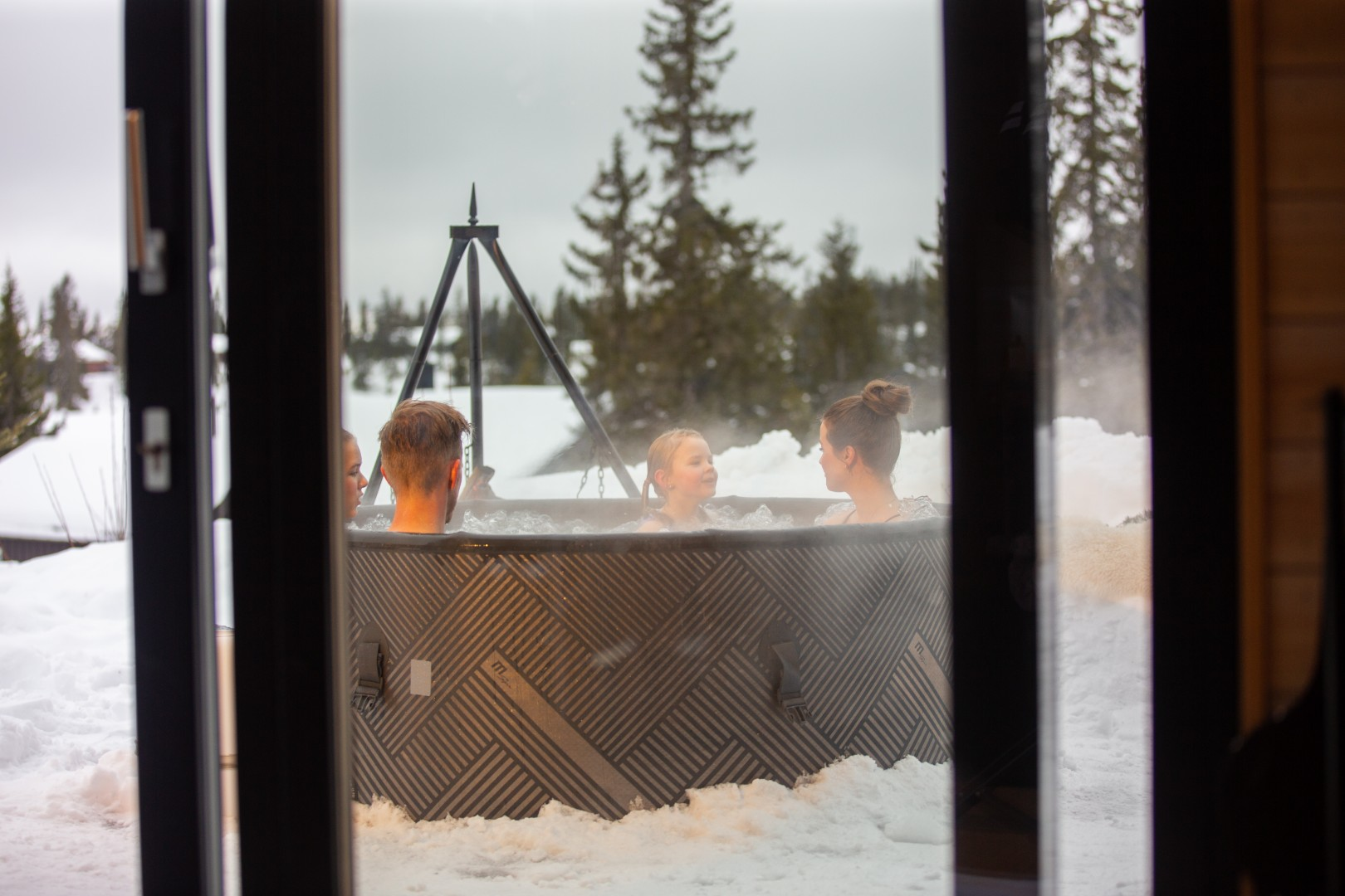 Menschen in einem Whirlpool im Freien, Schnee umgibt das Becken. Durch eine offene Tür betrachtet. Bäume und ein bewölkter Himmel sind im Hintergrund.