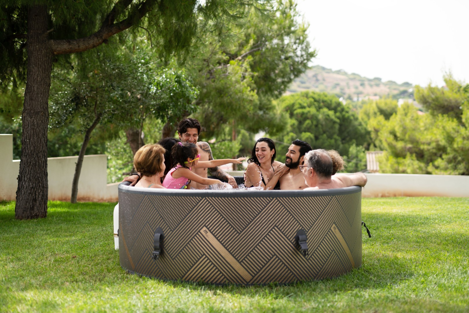 Eine Gruppe von Leuten in einem Whirlpool im Freien, umgeben von grünem Gras und Bäumen. Sie lächeln und interagieren.