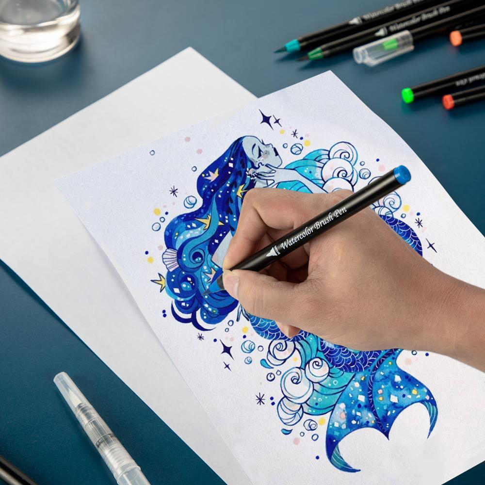 Una mano disegna una sirena blu con un pennarello acquerello. Altre penne e un bicchiere d'acqua sono sullo sfondo su una superficie blu.