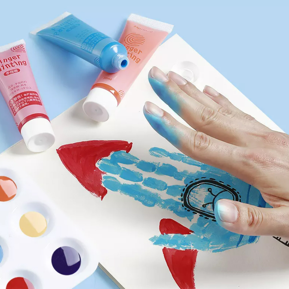 Una mano con pintura azul en los dedos creando una forma de cohete. También se ven tubos de pintura y una paleta. El fondo es azul claro.