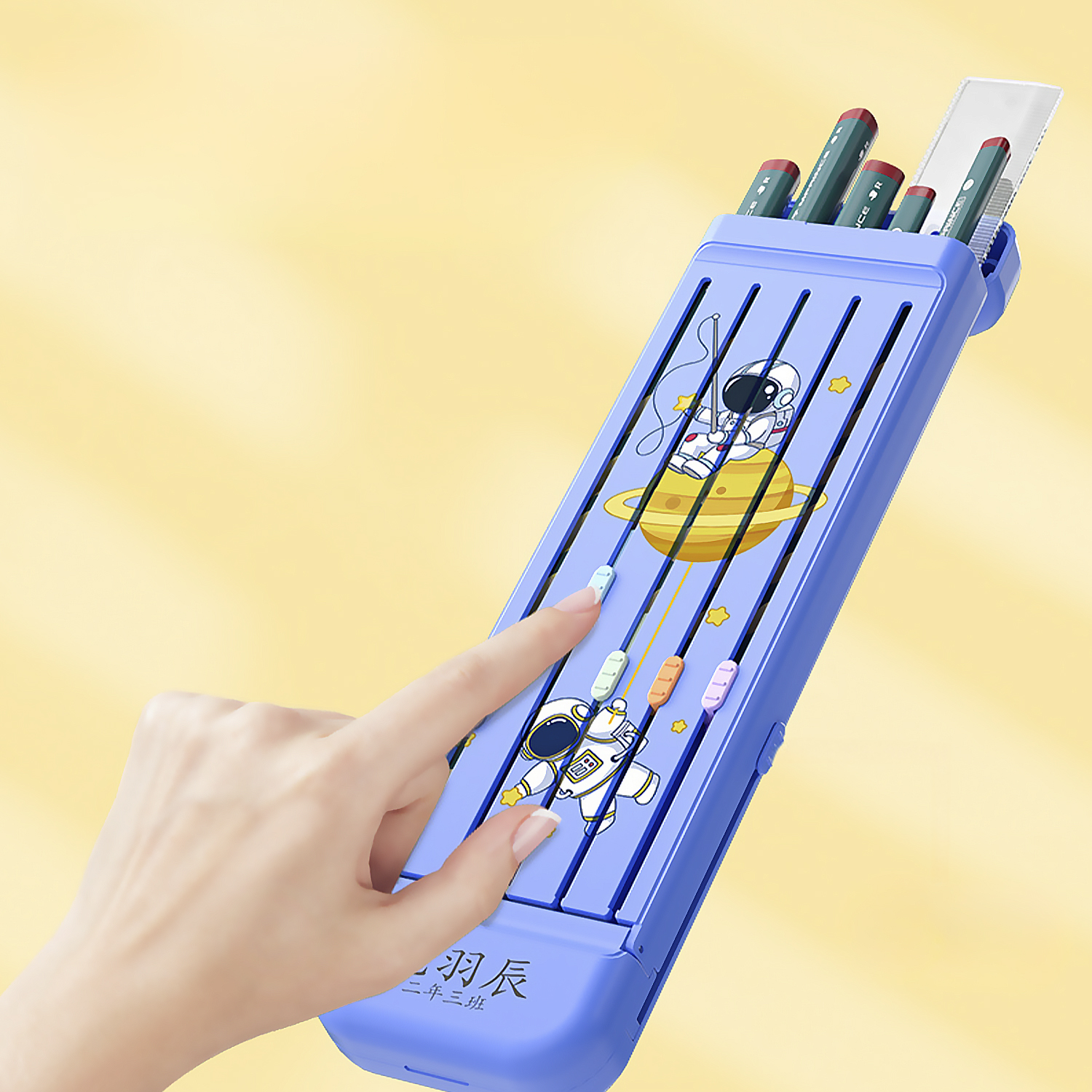 Una mano toca un estuche azul con ilustraciones espaciales. Se ven lápices dentro. El fondo es amarillo.