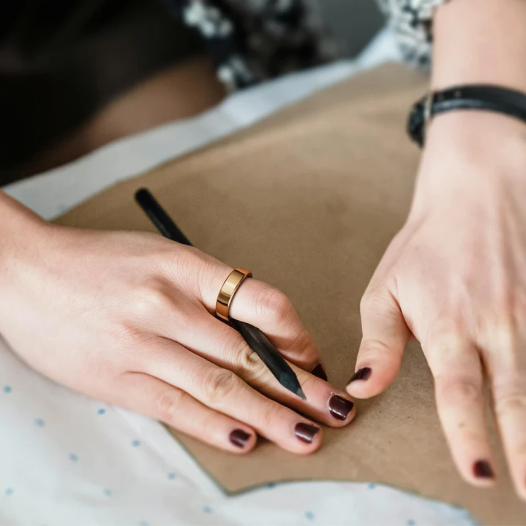 Eine Person zeichnet mit einem Bleistift auf braunem Papier. Die Person hat einen goldenen Ring und dunklen Nagellack.