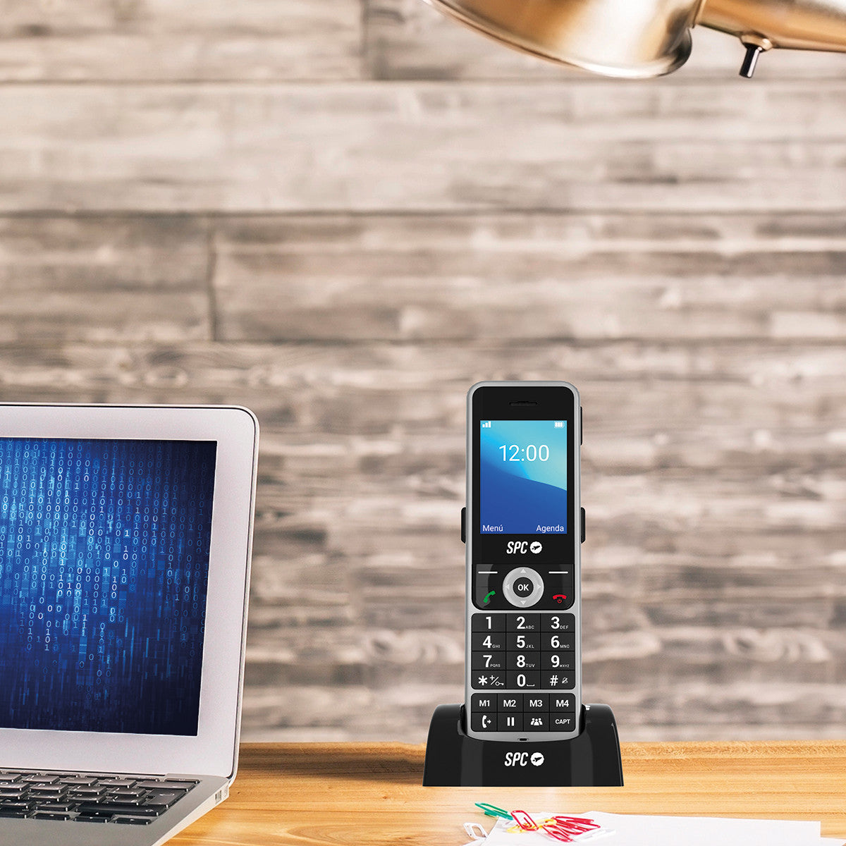 Un teléfono celular en una base de carga está sobre un escritorio. Una computadora portátil está a la izquierda. El fondo es una pared de madera.