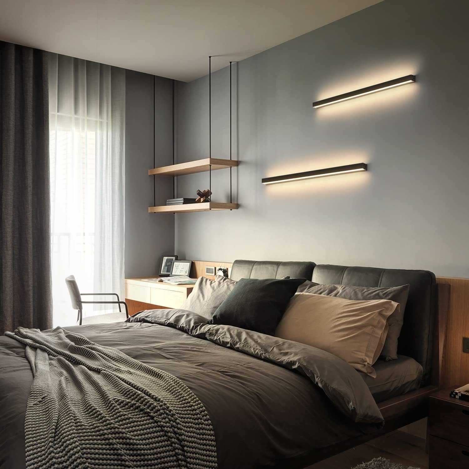 Un dormitorio moderno con cama, escritorio y estantes flotantes. Ropa de cama gris, luces de pared y una ventana con cortinas.