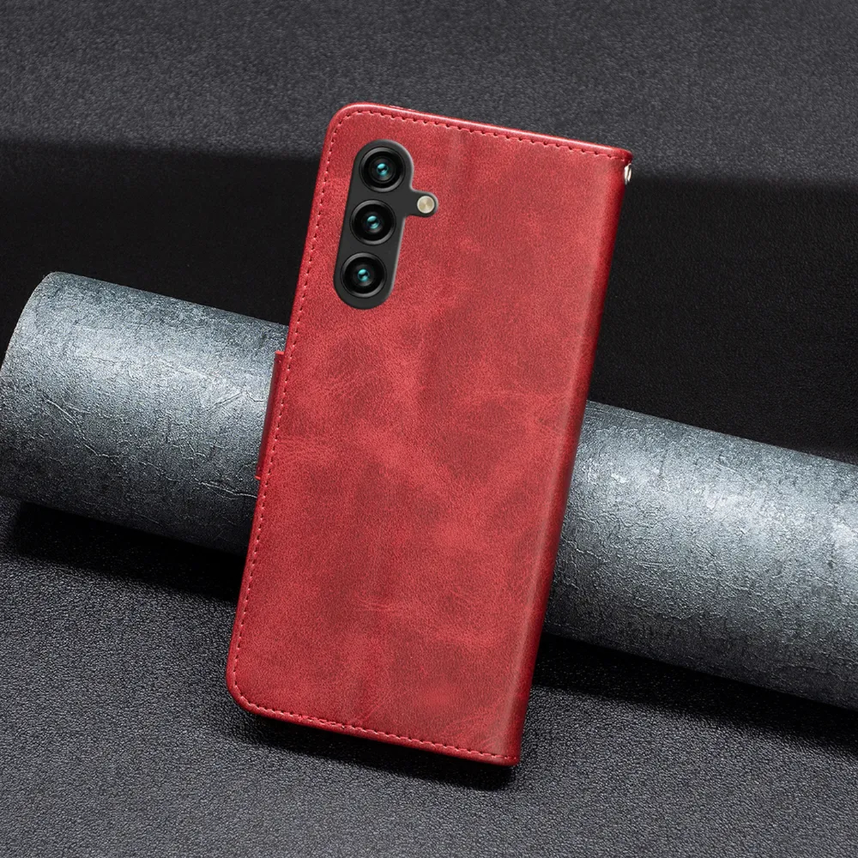 Een rode leren telefoonhoes met drie cameralenzen ligt op een getextureerd donker oppervlak met een cilindrisch object.