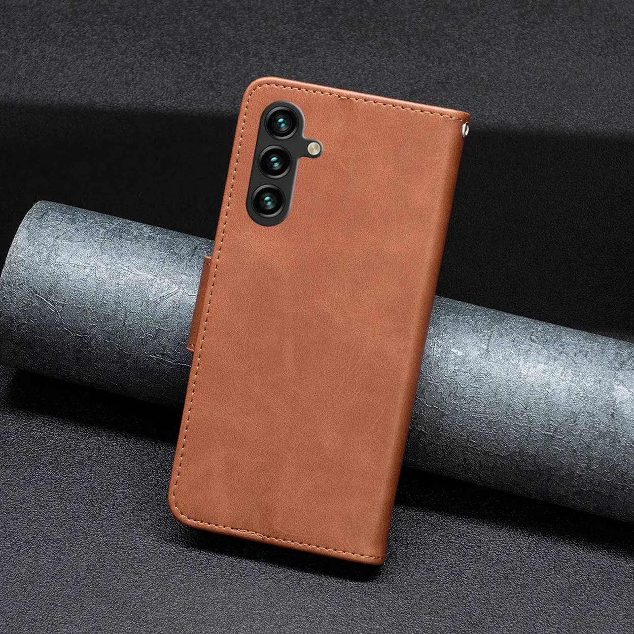 Bruine leren telefoonhoes met drie cameralenzen. Het ligt op een getextureerd donker oppervlak met een gerold grijs materiaal.