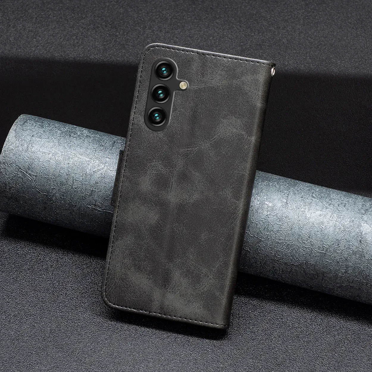 Een donkergrijze telefoonhoes ligt op een gestructureerd oppervlak, met een cilindrisch object op de achtergrond. De hoes heeft drie cameralenzen.