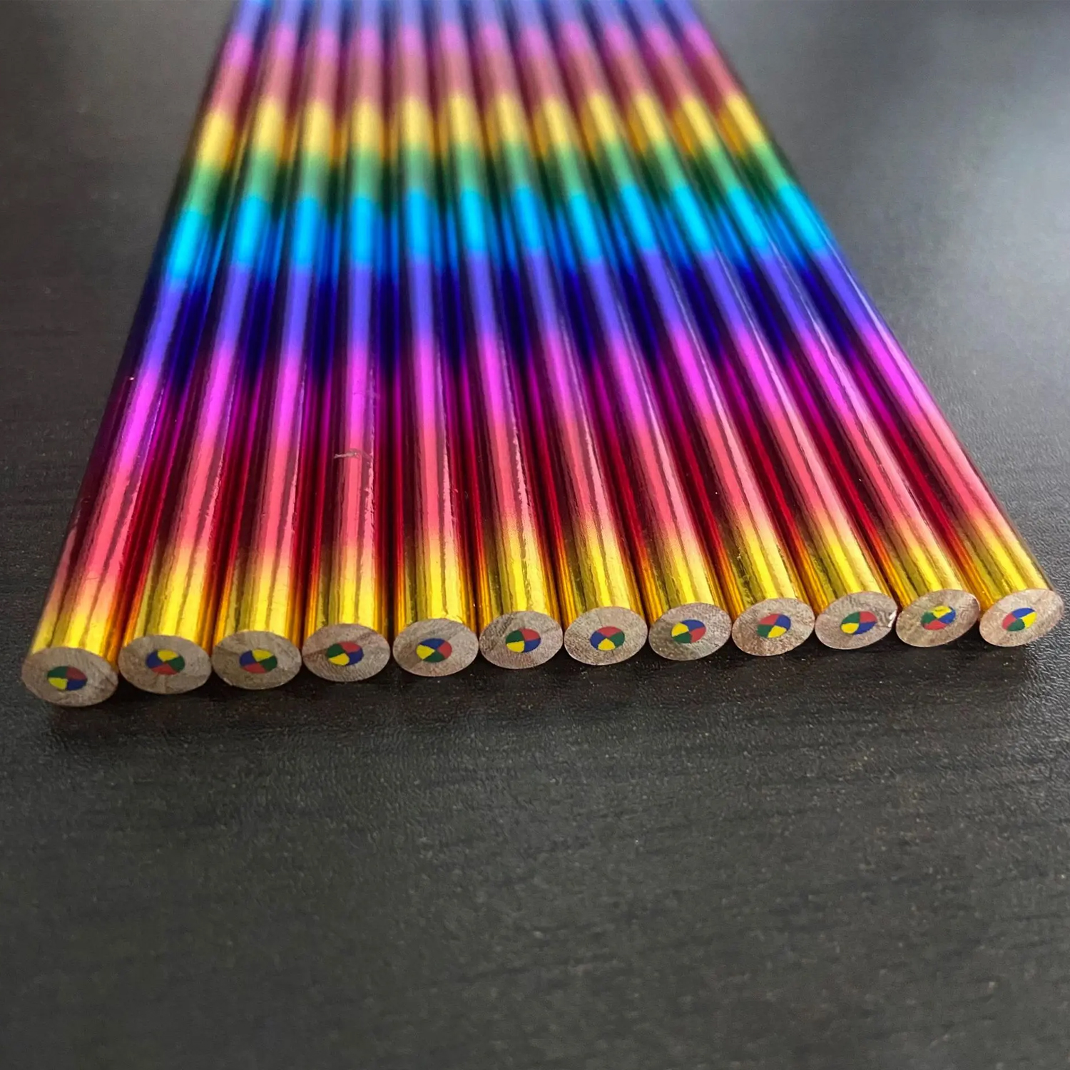 Primer plano de lápices de colores dispuestos en diagonal. Cada lápiz tiene un cuerpo arcoíris y una punta multicolor. El fondo es oscuro.