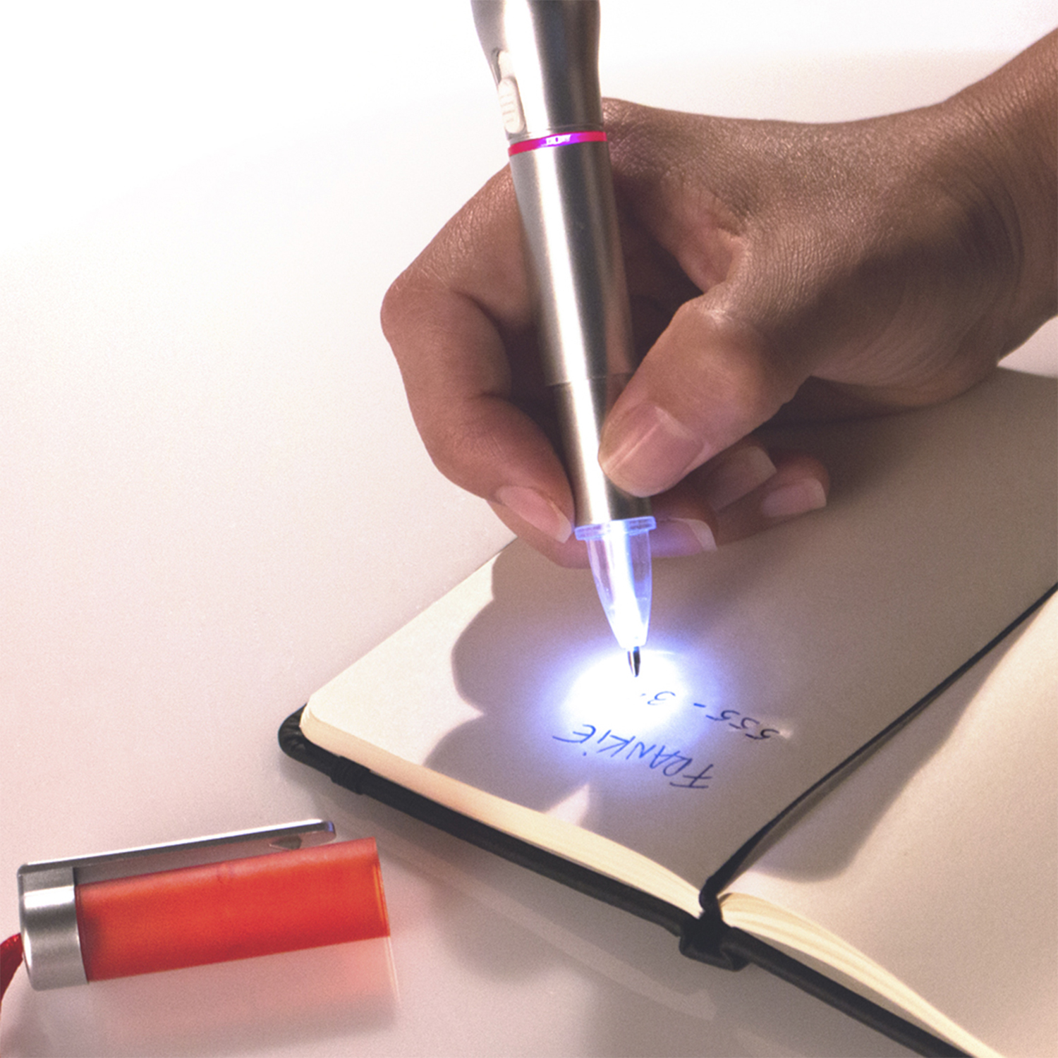 Eine Hand, die einen Stift mit leuchtender Spitze hält und auf ein offenes Notizbuch schreibt. Darunter liegt ein orangefarbenes Objekt mit roter Schnur.